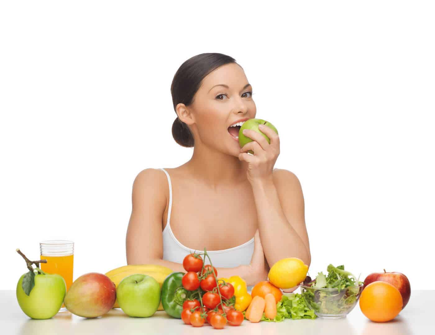 A woman eating an apple with fruits and vegetables in front of her.